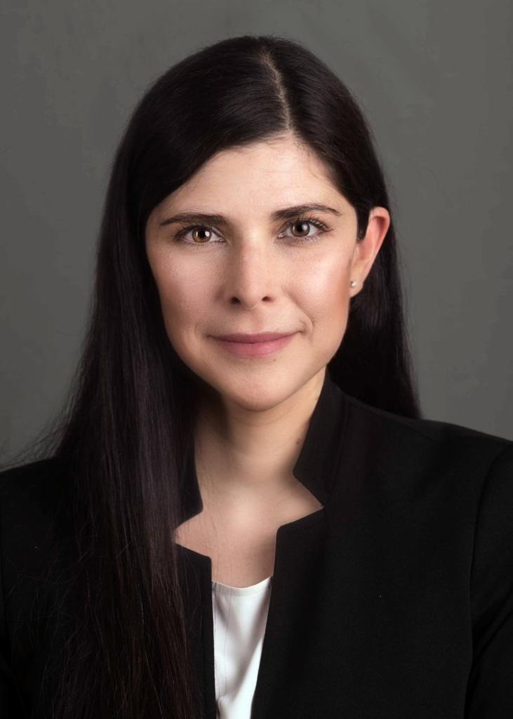 A professional headshot of a woman with long dark hair, wearing a black blazer over a white top, against a neutral background.
