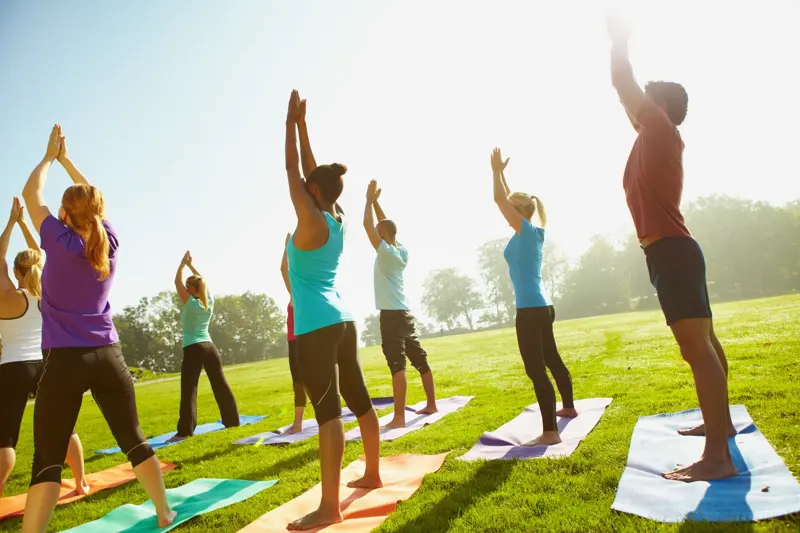 A racially diverse group of fit-looking people stretch as they face away from the camera at an outdoor yoga class held in a grassy field on a sunny day (iStock)