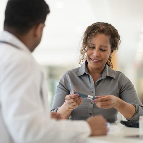 A male Indian doctor meets with a female patient. The woman is a mature adult of African descent. The two people are seated at a table. The patient is diabetic and is holding an insulin pen. The doctor is explaining how to use the device.