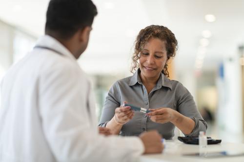 A male Indian doctor meets with a female patient. The woman is a mature adult of African descent. The two people are seated at a table. The patient is diabetic and is holding an insulin pen. The doctor is explaining how to use the device.