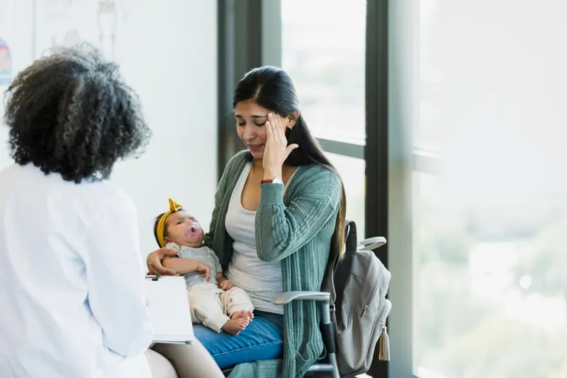 The mid adult woman with a baby touches her head and shows distress as she describes her pain to the unrecognizable mature adult female specialist.