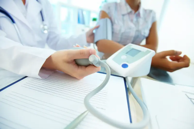 Close-up photo (heads not shown) of a person wearing a white lab coat and stethoscope gripping the bulb of a blood pressure measuring device (tonometer) while the other end is attached to a patient's arm (Shutterstock 113625799)