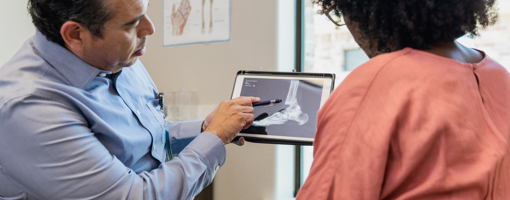 The mature adult orthopedist uses a pen to point out the injury site on the x-ray to the unrecognizable female patient.