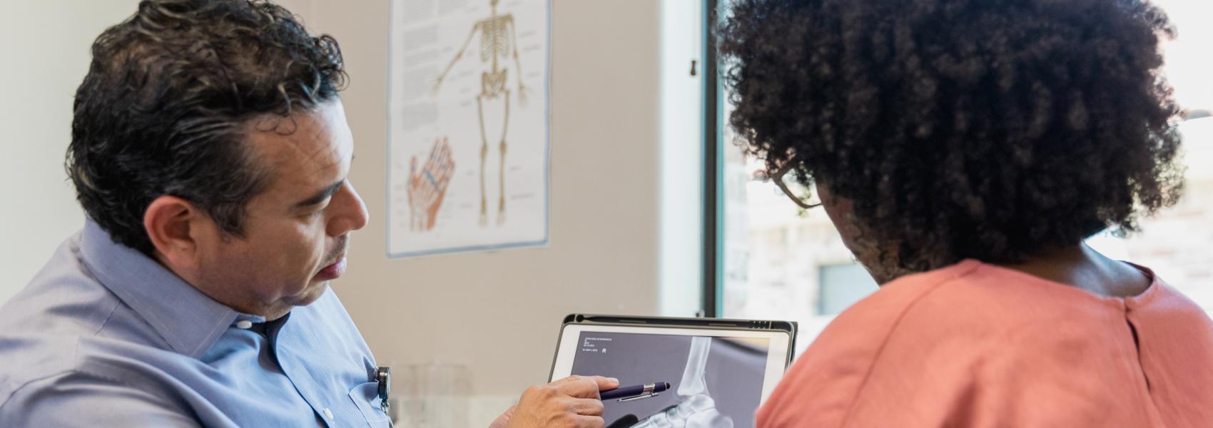 The mature adult orthopedist uses a pen to point out the injury site on the x-ray to the unrecognizable female patient.