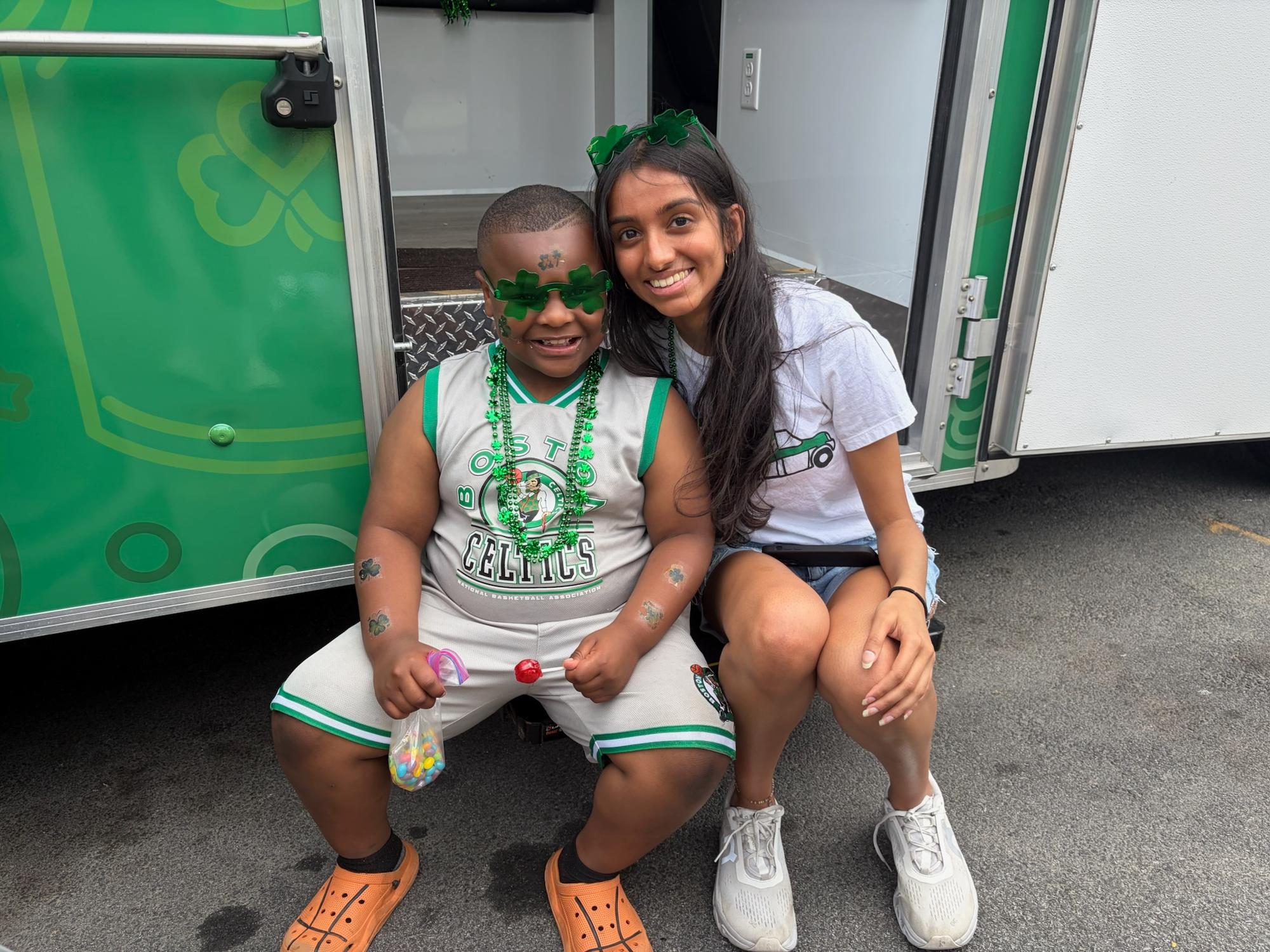 Event photo from the Dorchester Day Parade (June 2026): BMC staff member sitting with a child (both dressed in Boston Celtics swag) in front of the BMC Curbside Care postpartum and newborn health mobile van