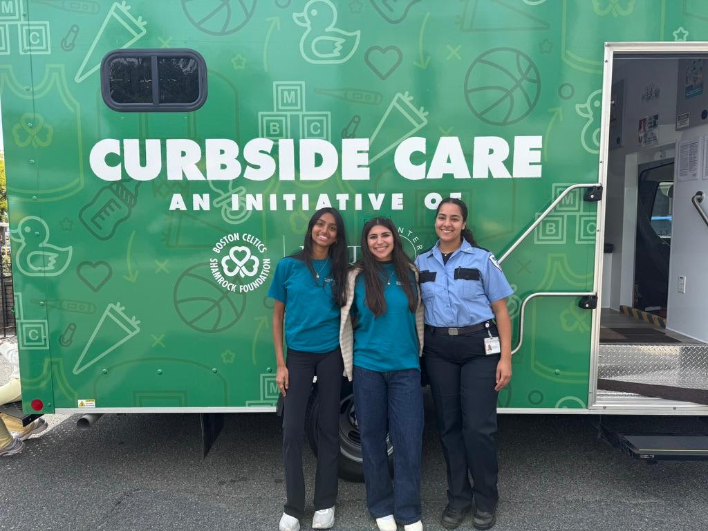 BMC Community Wellness Advocate Sam Jangala, volunteer Emma Santos, and Brewster Ambulance Services driver Fior Pena pose in front of the Curbside Care van at the September 2026 Live Well Boston! Community Health and Wellness Block Party in Dorchester, hosted by MelanIN Medicine, Inc.