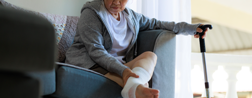 In a somber scene, a senior Asian woman is sitting on a sofa, looking sad. She holds a walking stick, and there's a bandage on her foot, indicating that she may be dealing with an injury or health issue.