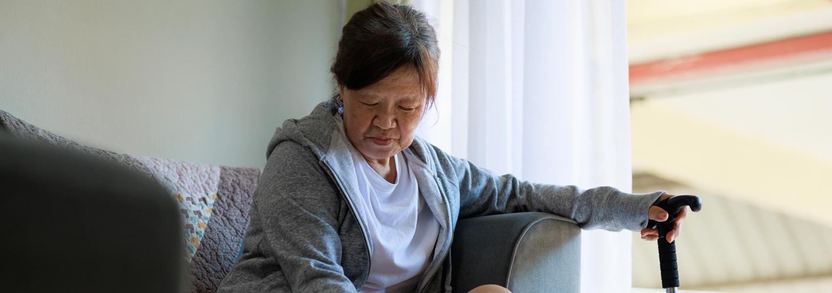 In a somber scene, a senior Asian woman is sitting on a sofa, looking sad. She holds a walking stick, and there's a bandage on her foot, indicating that she may be dealing with an injury or health issue.