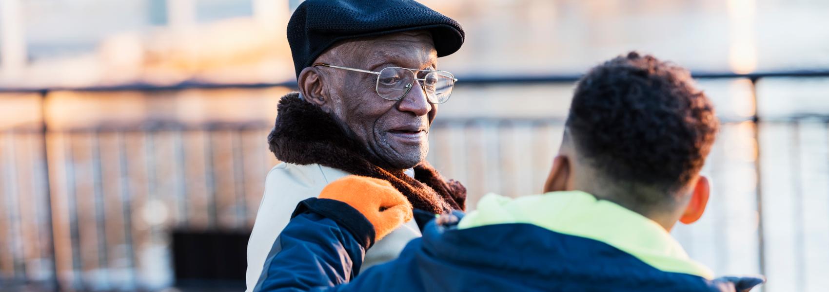 An African-American boy, 10 years old, sitting by a city waterfront with his great grandfather, a senior man in his 70s, who is looking at him affectionately, smiling. The child's back is to the camera. It is autumn or winter so they are wearing warm clothing.