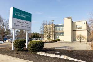 Exterior of the BMC Health Primary Care (Whitman) building (a modern beige brick building with a glass-walled entryway and stairwell), angled so that the adjacent parking area and the BMC Health System sign are visible in the foreground. The office is located at 312 Bedford Street, Whitman, MA 02382.
