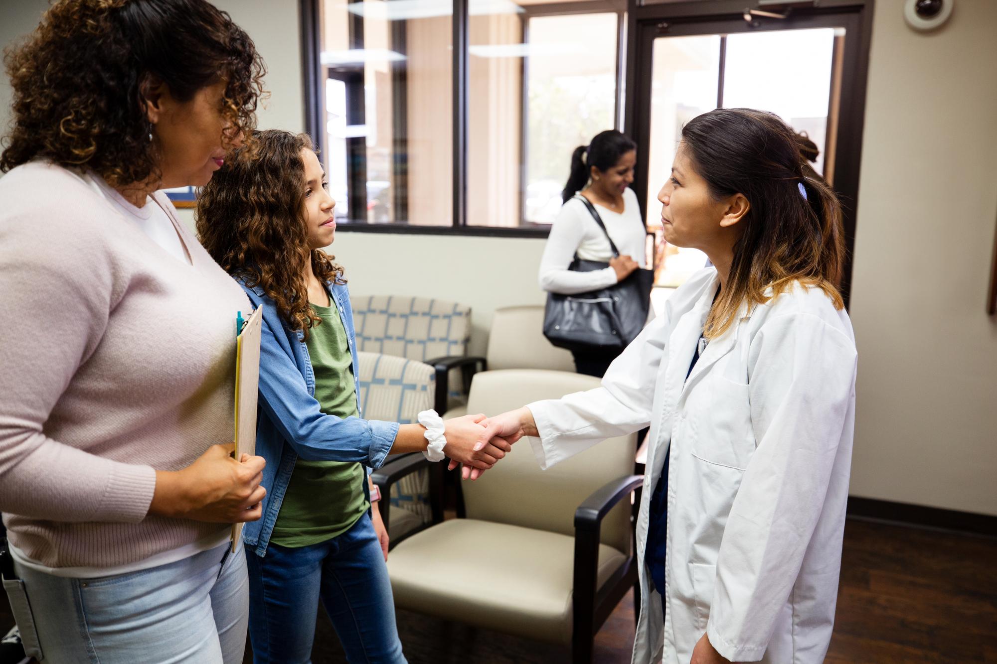 Friendly mid adult female doctor greets a preteen girl and the girl's mom. The doctor shake hands with the girl as her mom looks on.