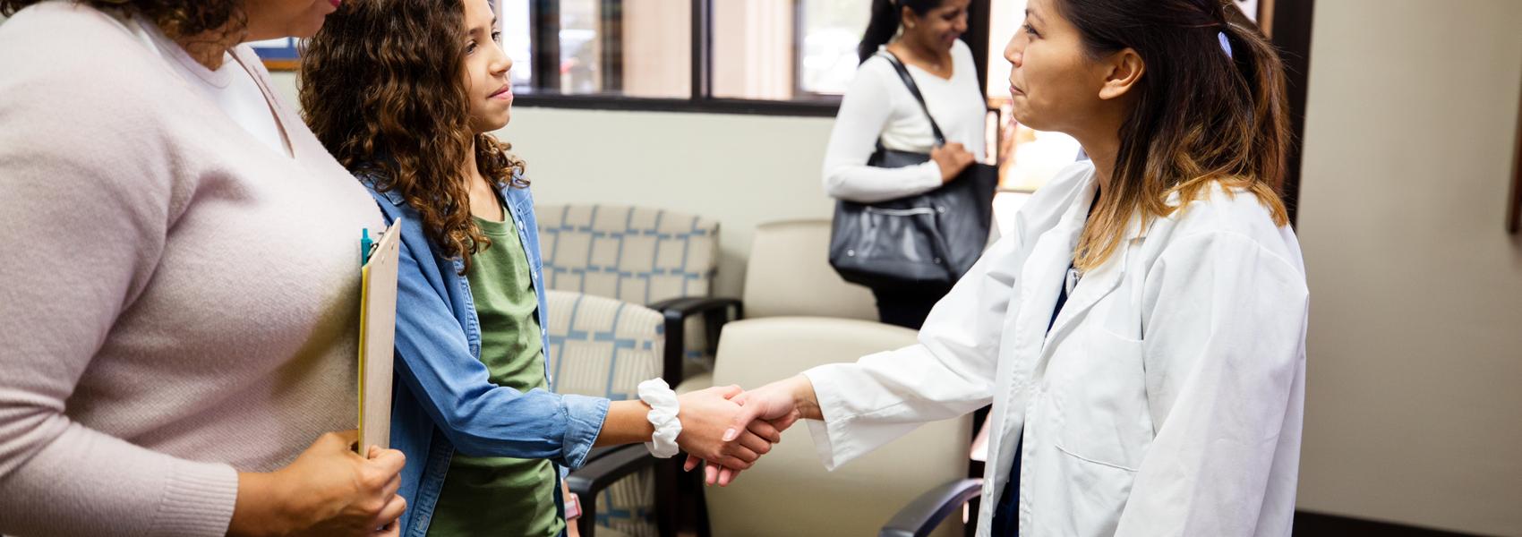 Friendly mid adult female doctor greets a preteen girl and the girl's mom. The doctor shake hands with the girl as her mom looks on.