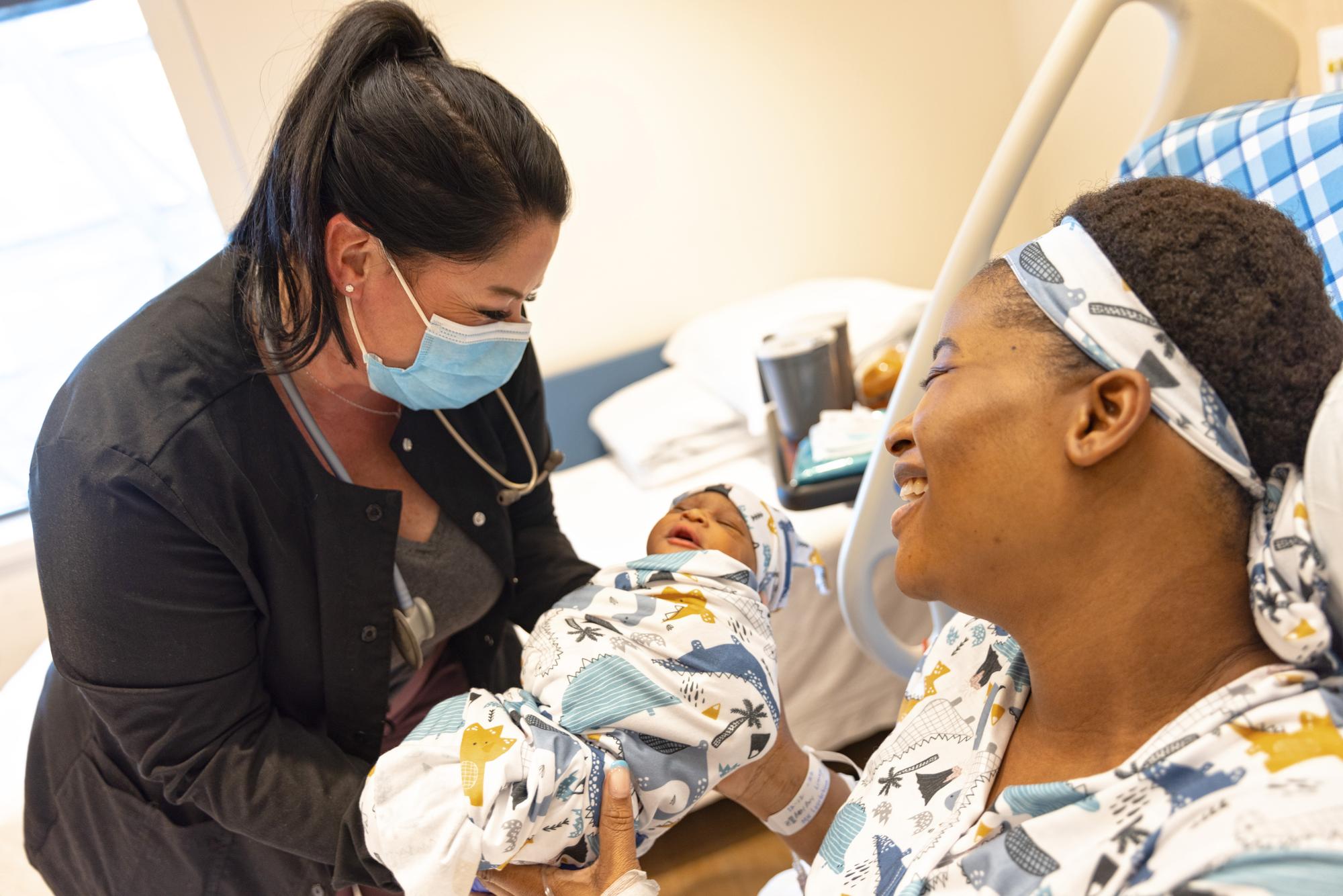 A labor and delivery nurse hands a swaddled newborn to a smiling new mother