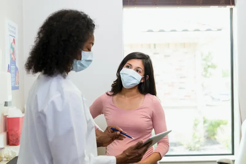 The mid adult woman and the mature adult female obstetrician wear protective masks due to the coronavirus epidemic.  The patient listen to the doctor.