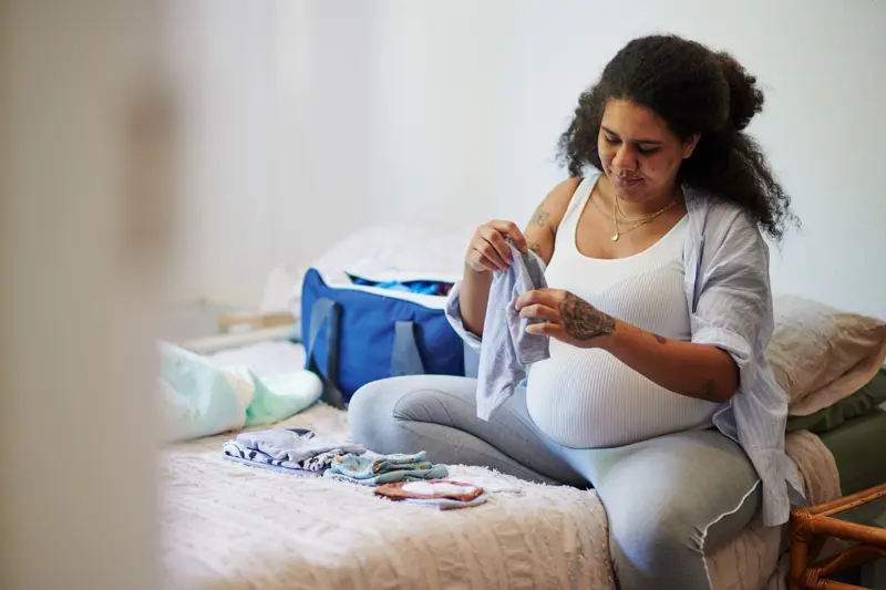 Pregnant Black woman smiles to herself as she folds newborn clothing while sitting on a bed in a bedroom. An open duffel bag is behind her on the bed.