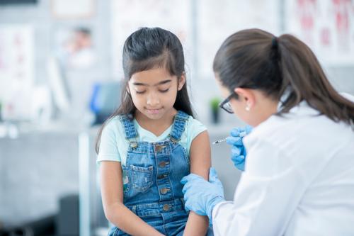 A little girl gets a vaccination from her doctor. She looks down as she wears a brave face. Her doctor is holding her arm.
