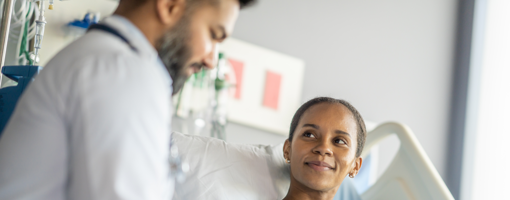 A woman sits up in a hospital bed wearing a gown as she talks with her male doctor about her surgery.  The doctor is taking notes electronically on a tablet as the two talk about the procedure.