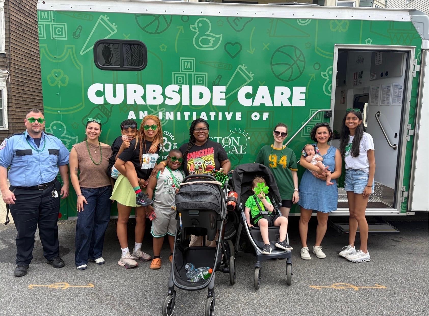 Event photo from the Dorchester Day Parade (June 2026): BMC staff and community members standing/sitting in front of the BMC Curbside Care postpartum and newborn health mobile van