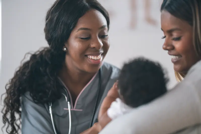 Close up shot of a young black nurse meeting with a patient. The patient is a baby. The child is being held by her mother. The smiling medical professional is leaning in close to examine the baby.